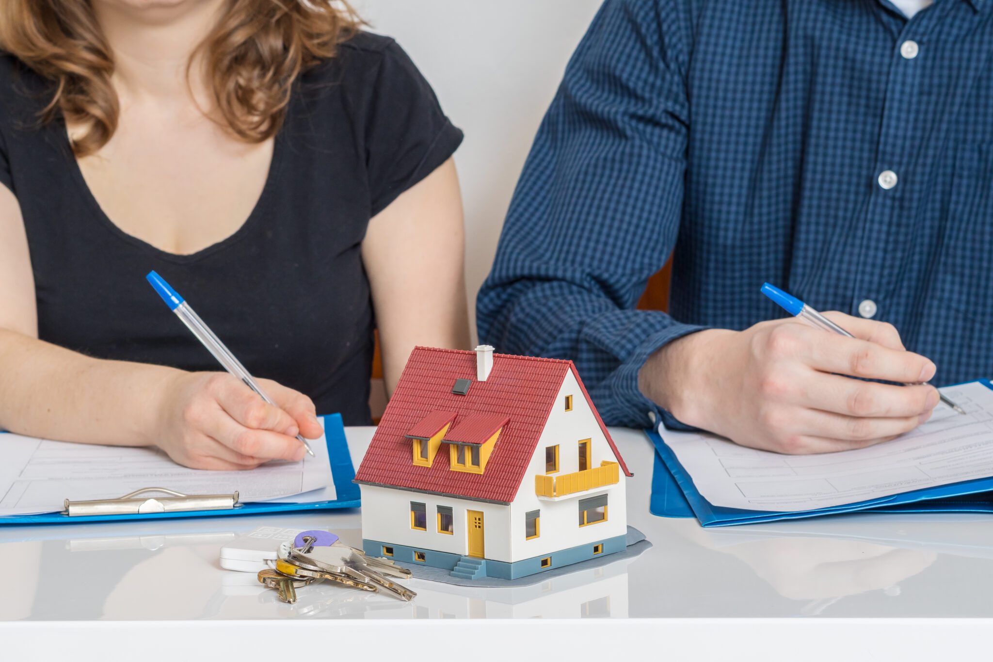A couple signing documents with a house model and keys on the table.
