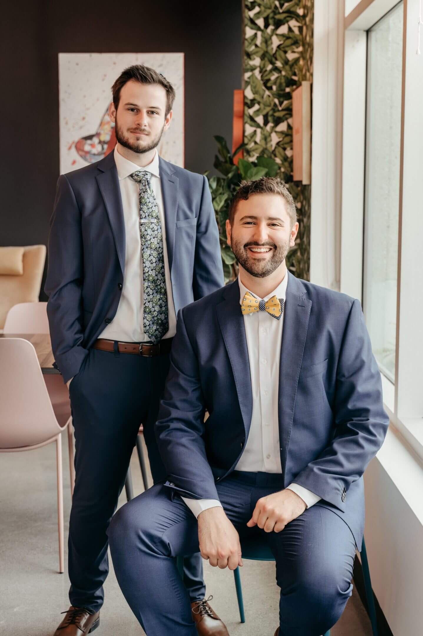 Two men in suits posing indoors, one seated and one standing.