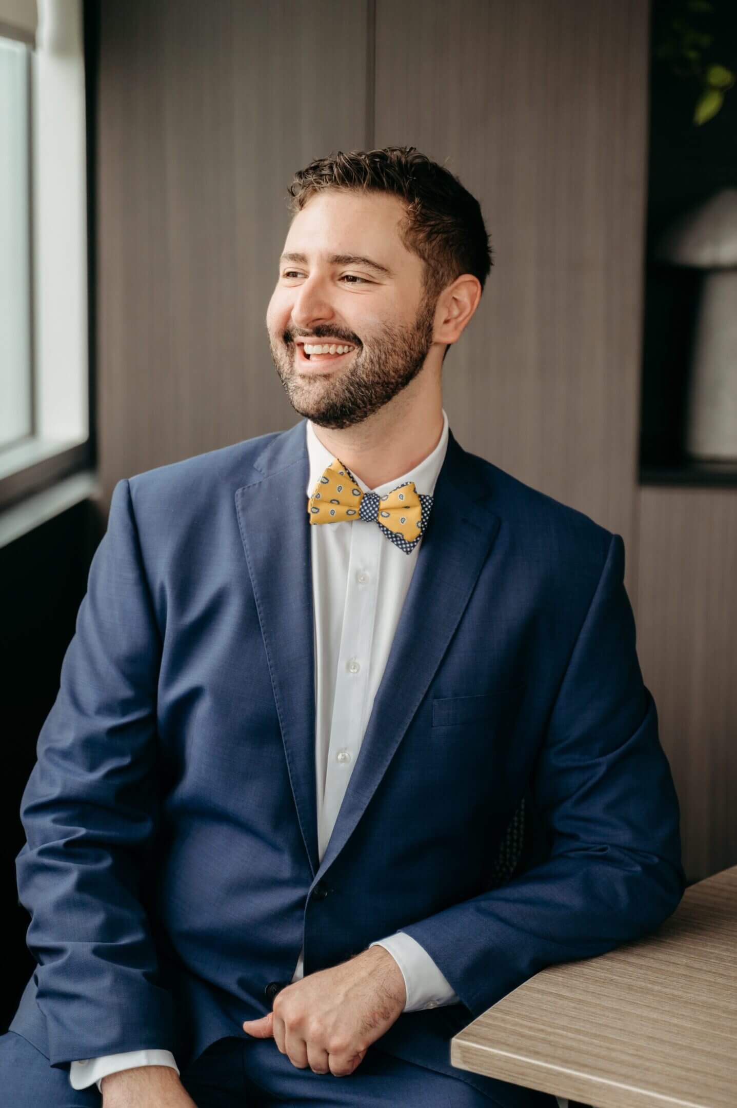 A smiling man in a blue suit and yellow bow tie near a window.