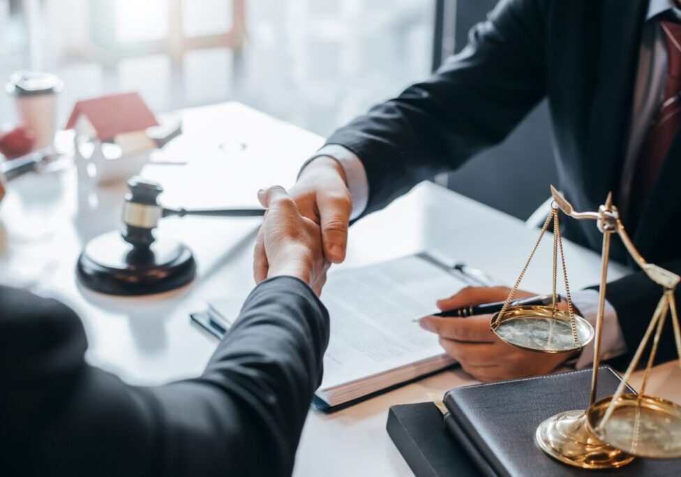 Two people shaking hands over a legal desk with scales and documents.