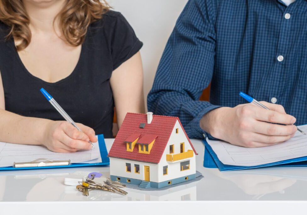 A couple signing documents with a house model and keys on the table.
